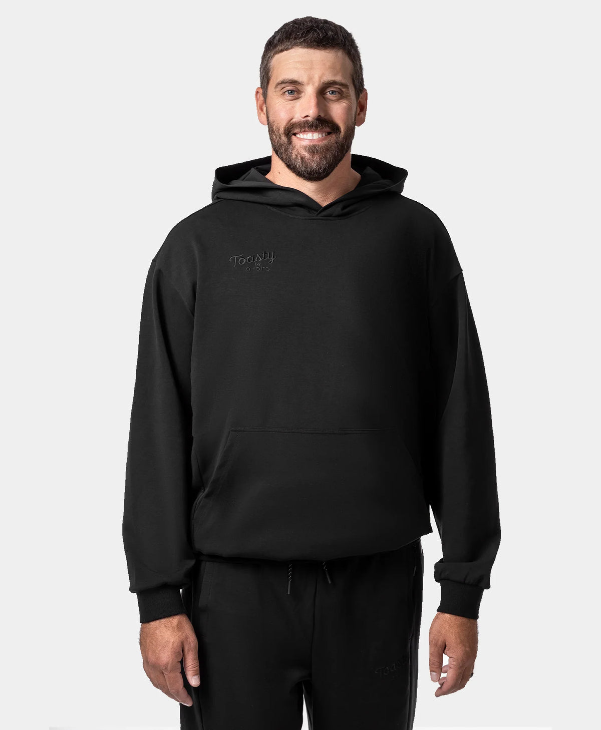 A smiling man wearing a black Toasty Unisex Heated Pullover Hoodie and matching sweatpants with a subtle tonal embossed "Toasty by ororo" logo on the left chest, photographed against a plain light gray studio background.