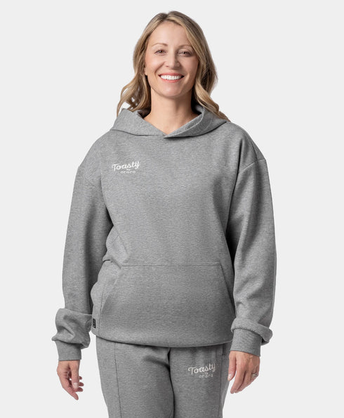 A smiling woman wearing a flecking gray Toasty Unisex Heated Pullover Hoodie and matching sweatpants with a small white "Toasty by ororo" logo printed on the left chest and upper thigh, photographed against a plain off-white studio background. ,view 1